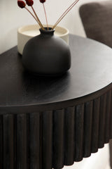 Close-up of a black round side table with ribbed vertical wood detailing and matte black ceramic vase holding dried red-brown floral stems.