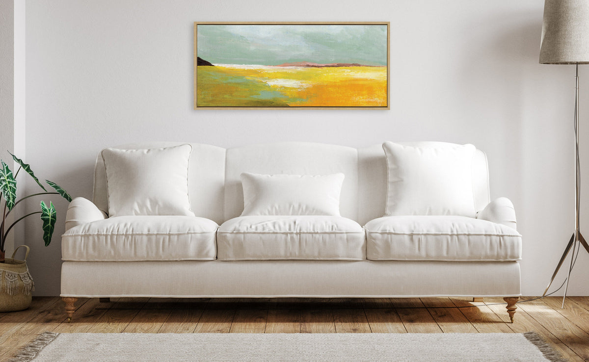 Framed rectangular canvas above white sofa, depicting abstract landscape with sky-blue, golden yellow, and green shades on a minimalist off-white wall.