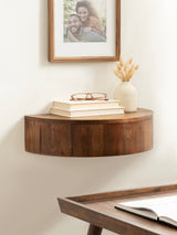 Half-round floating wood side table mounted on a light wall, holding stacked books with glasses and a small beige vase with dried grasses, below a framed photo.
