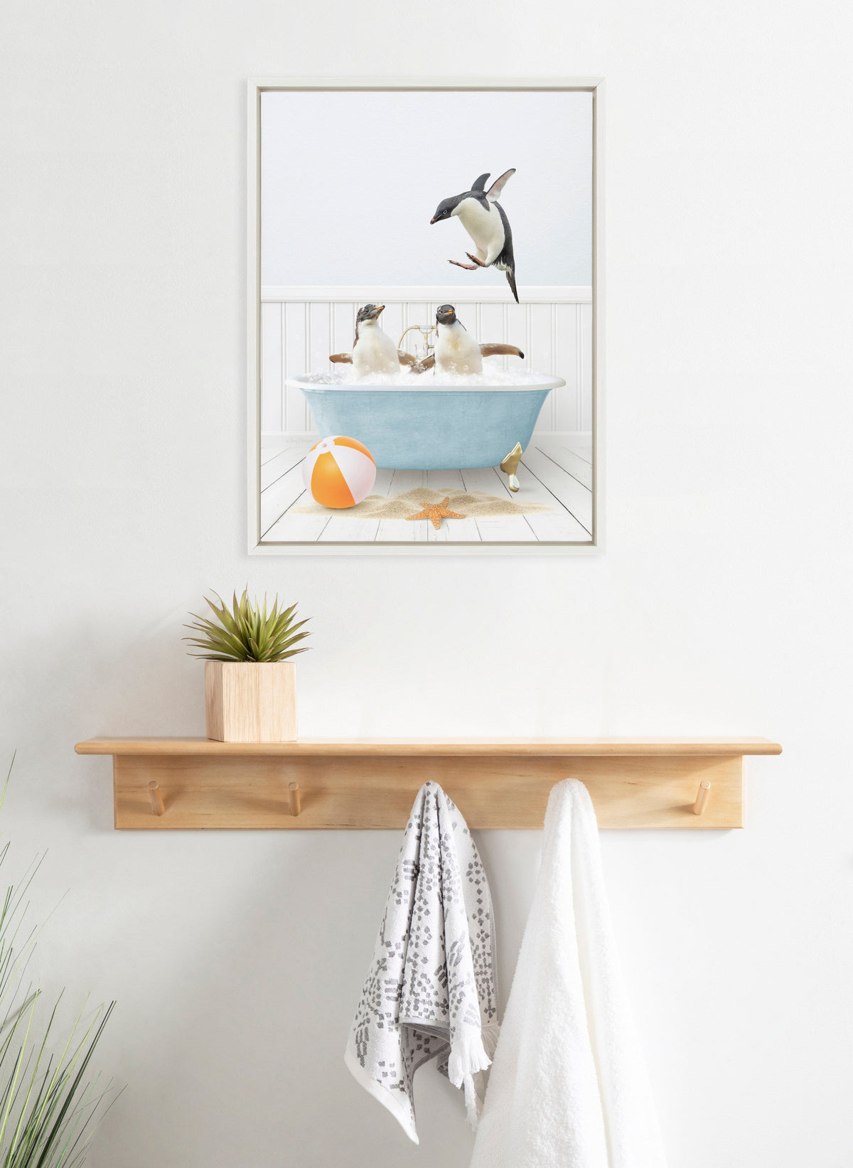Framed canvas artwork depicting three penguins in and around a blue bathtub on a deck, with a striped beach ball and starfish on the floor.