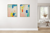 Two abstract paintings with blue, yellow, pink, red, and teal brushstrokes, framed in light wood, hanging side by side on a beige wall above white baseboard molding.