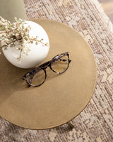 Round golden metal side table top with textured surface holding a white vase with dried flowers and a pair of tortoiseshell eyeglasses, on a patterned rug.