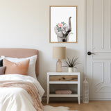 Framed print of a gray elephant with a raised trunk adorned with pink and white flowers hanging above a white nightstand in a softly lit bedroom.