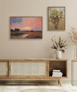 A warm-toned living room wall featuring two framed paintings: a horizontal landscape with a pink sunset over fields and a vertical floral still life with colorful flowers and butterflies.