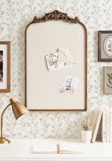 Rectangular beige fabric pinboard with ornate dark bronze arch frame, displaying pinned envelopes and photos, mounted on floral wallpaper above white desk with gold lamp.