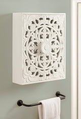 White carved wood wall cabinet with ornate floral and scrollwork patterns, mounted on a light gray wall above a black towel bar holding a white towel.