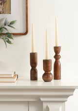 Three dark wood taper candle holders with varying shapes hold white taper candles on a white mantel beneath a framed wall art and green leafy plant.
