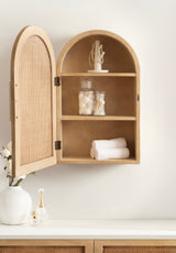 Light wood arched wall cabinet with open mesh door, three shelves holding cotton balls, swabs, rolled towels, and a jewelry stand against a white wall.