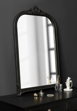 Arched black wall mirror with ornate carved frame and beaded edge, resting on a dark wooden surface reflecting window light, with small glass and ceramic decorative items nearby.