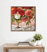 Framed close-up of a floral arrangement featuring red, white, and peach flowers with green leaves in a tarnished brass goblet, displayed on a white cloth background.