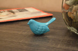 Small turquoise bird figurine with carved feather details resting on dark wood surface near a glass container filled with stones and a blurred magazine.
