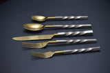 Set of five golden flatware pieces with textured silver-tone handles, including two spoons, one knife, and two forks, arranged horizontally on a dark surface.