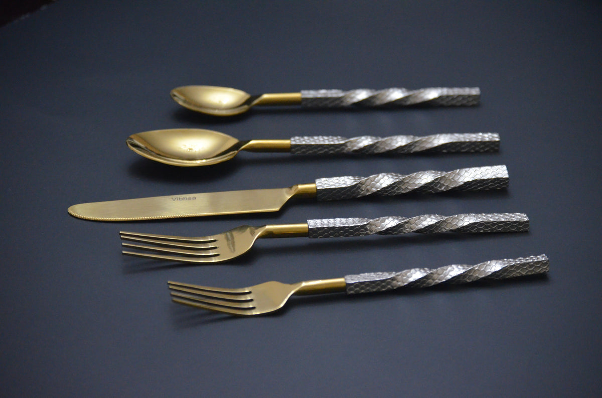 Set of five golden flatware pieces with textured silver-tone handles, including two spoons, one knife, and two forks, arranged horizontally on a dark surface.