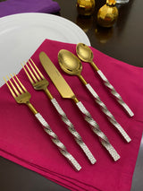Six-piece golden flatware set with twisted textured silver handles arranged on a magenta napkin over a dark wooden table with two small golden decorative items in the background.