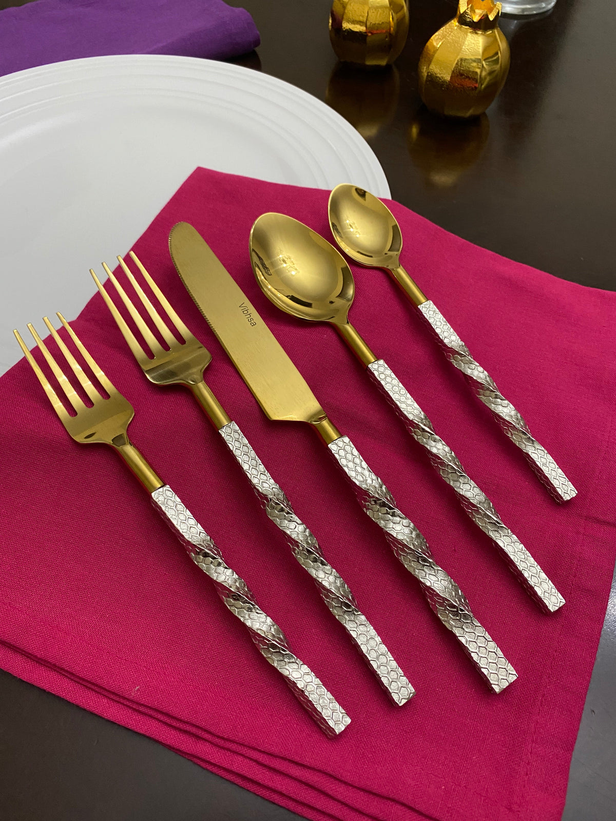 Six-piece golden flatware set with twisted textured silver handles arranged on a magenta napkin over a dark wooden table with two small golden decorative items in the background.