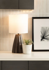 Table lamp with a dark brown scalloped base and a white cylindrical lampshade on a white surface, next to a small white pot with a green air plant and a black-framed abstract photo.