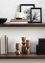 Three bronze-colored tapered candle holders with distinct curvy shapes arranged on a wooden shelf below a shelf holding neutral-toned books and two black-framed floral prints.