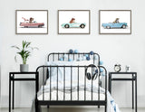 Three framed artworks of cartoon dinosaurs driving vintage cars in pink, light blue, and blue, hanging above a black metal bed with blue and white bedding and side tables.