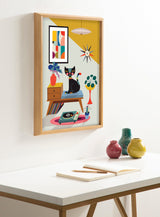 Framed colorful print showing a black cat on a mid-century modern wood side table with a record player, geometric wall art, starburst clock, and plants in a mustard and light blue room.