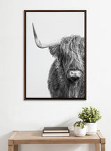 Black and white close-up portrait of a Highland cow with long shaggy hair partially obscuring its eyes, one prominent curved horn visible, framed and hung above a wooden table.