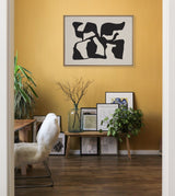 Black abstract shapes forming an irregular composition on a tan canvas, framed and hung on a mustard yellow wall above a wooden bench with plants and framed art pieces beneath.
