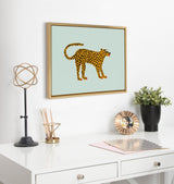 Framed canvas print of a stylized spotted cat with a curled tail on pale blue background, hanging above a white desk with decorative objects and a potted plant.