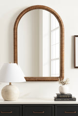 Arched wall mirror with a textured brown beaded frame reflecting a bright window, hung above a black cabinet with a round white lamp and a stack of books topped with a small white vase.