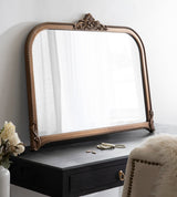 Arched wall mirror with bronze ornamental frame featuring rope-like and floral detailing, resting on a dark wood surface beside a beige studded chair and flowers.