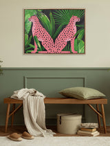 Framed canvas artwork featuring two pink cheetahs with black spots sitting back-to-back against a green tropical leaf background, mounted on a beige wall above a wooden bench.