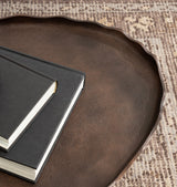 Oval metal tray with a dark brown textured surface and wavy edge, holding two black hardcover books with cream-colored pages, on a patterned rug.