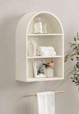Cream-colored arch-shaped wall shelf with three compartments holding cotton balls, rolled towels, soap dispenser, framed photo, and small vase with flowers.