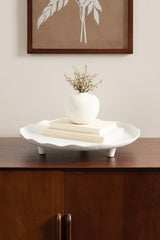 White scalloped ceramic tray with short rounded legs, holding a small white vase with dried flowers and two cream-colored books on a wooden sideboard under a framed leaf artwork.