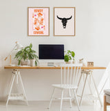 Minimalist workspace with natural wood sawhorse desk, white chair, green potted plants, and two framed artworks featuring an orange cowgirl playing card and a black bull skull on cream backgrounds.