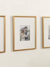 Three vertical gold photo frames with small bead details, each containing a portrait photo, mounted on a cream-colored wall in a row.