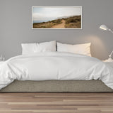 Framed landscape canvas of a coastal path winding through green and brown shrubs under a cloudy sky, hung above a bed with white bedding and a gray frame.