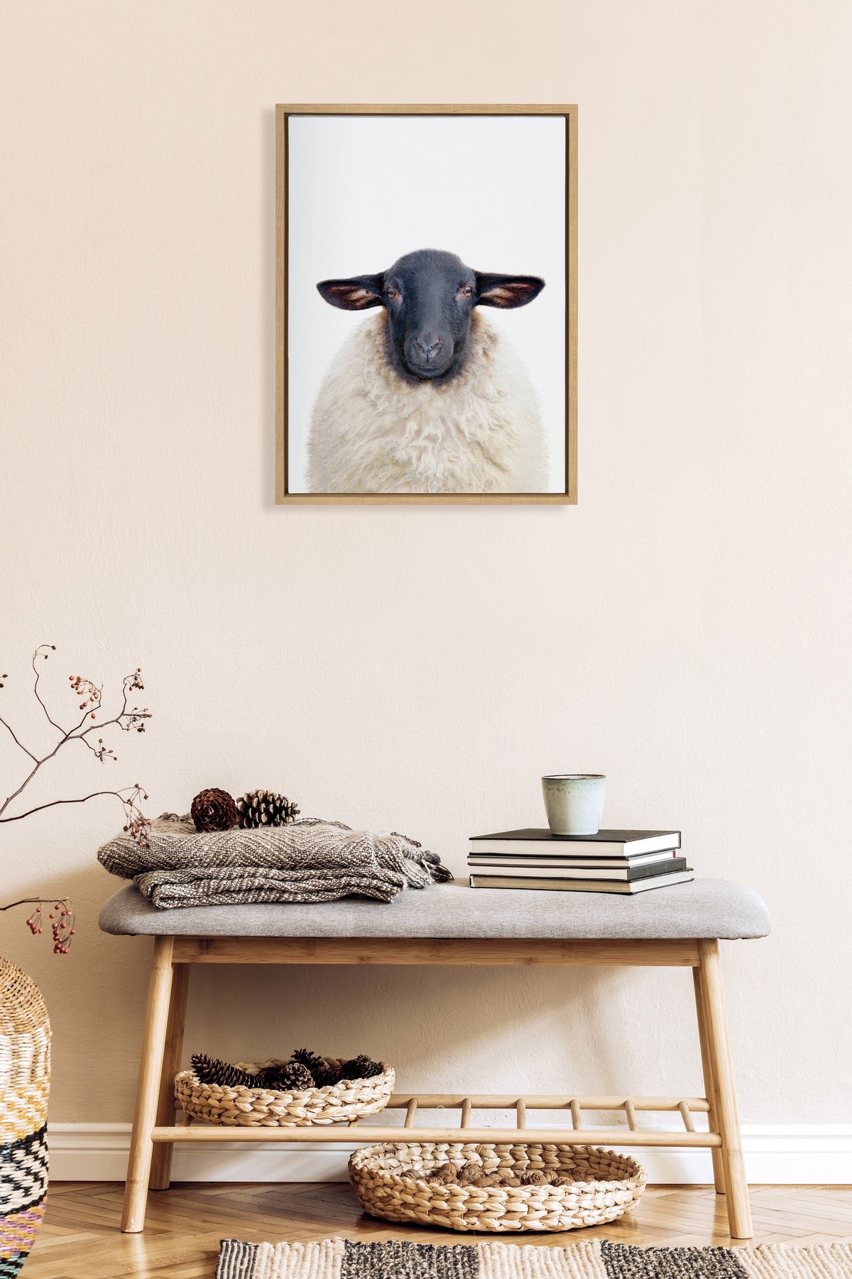 Framed portrait of a white sheep with a black face centered on a light wall above a wooden bench holding folded blankets, books, and a cup, with wicker baskets below.