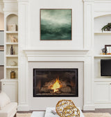 Square abstract canvas with tonal sage green gradients and soft textures, framed in dark wood, hanging above a lit fireplace in a bright white living room.