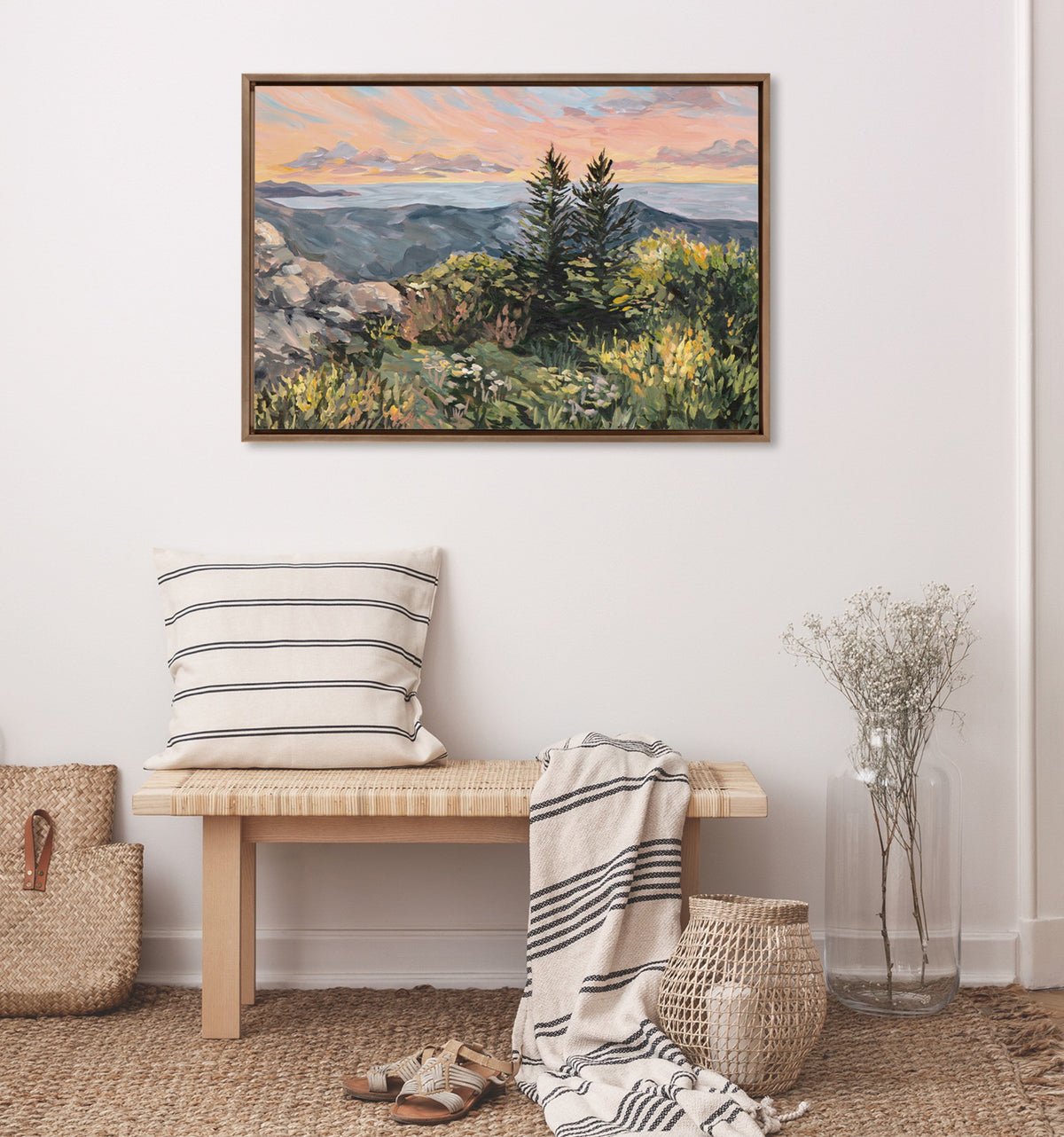 Framed landscape painting with pine trees and rocky foreground under a pastel sunrise sky, hung above a wooden bench with striped cushion and draped throw.