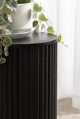 Close-up of a black cylindrical side table with vertical grooves, topped with a white planter holding a plant with round, variegated green leaves against a sheer white curtain.