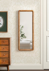 Tall rectangular wall mirror with a rounded-edge natural wood frame featuring a beaded texture, reflecting a white couch and green plant leaves.