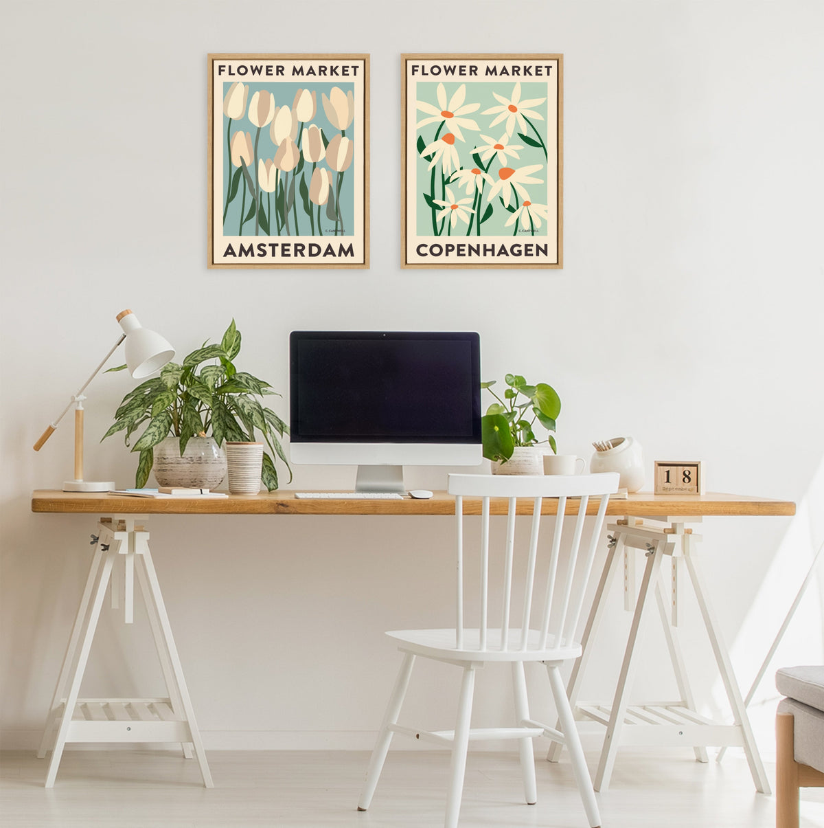 Minimalist workspace with a wooden top desk and white legs, featuring two framed flower market prints labeled Amsterdam and Copenhagen on wall above.