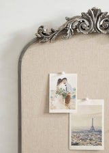 Close-up of an ornate silver arch frame with intricate scrollwork, enclosing a beige fabric pinboard holding two pinned photographs, one of a couple and one of the Eiffel Tower.