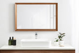 Rectangular wall mirror with a medium brown wooden frame reflecting light and shadows, mounted above a white rectangular sink on a white countertop with green plants and dark bottles.