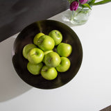 Dark ribbed metal bowl holding several green apples, shown from above on a white surface with a glass vase of purple tulips nearby