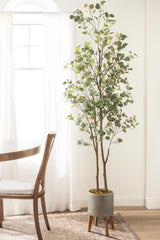 Tall faux eucalyptus tree with round green leaves in a textured gray pot on wooden legs, placed next to a wooden chair and round table by a sheer-curtained window.