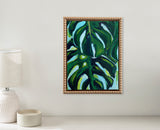 Framed canvas painting of dark green Monstera leaves with highlights of lighter green and blue, displayed on a beige wall above a white lamp and candle.