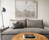 Rectangular black pillow with indigo and white matchstick pattern on gray three-seat sofa, beside tall black floor lamp and abstract white-on-beige framed artwork.
