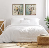 Two framed botanical paintings of vases with leaves and flowers in muted green, beige, and yellow hues hanging side by side above a white bedding set in a minimalist bedroom.