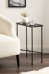 Slim black metal side table with a rectangular top holding a small white vase and a clear glass of water, positioned next to a cream upholstered armchair on dark wood floor.