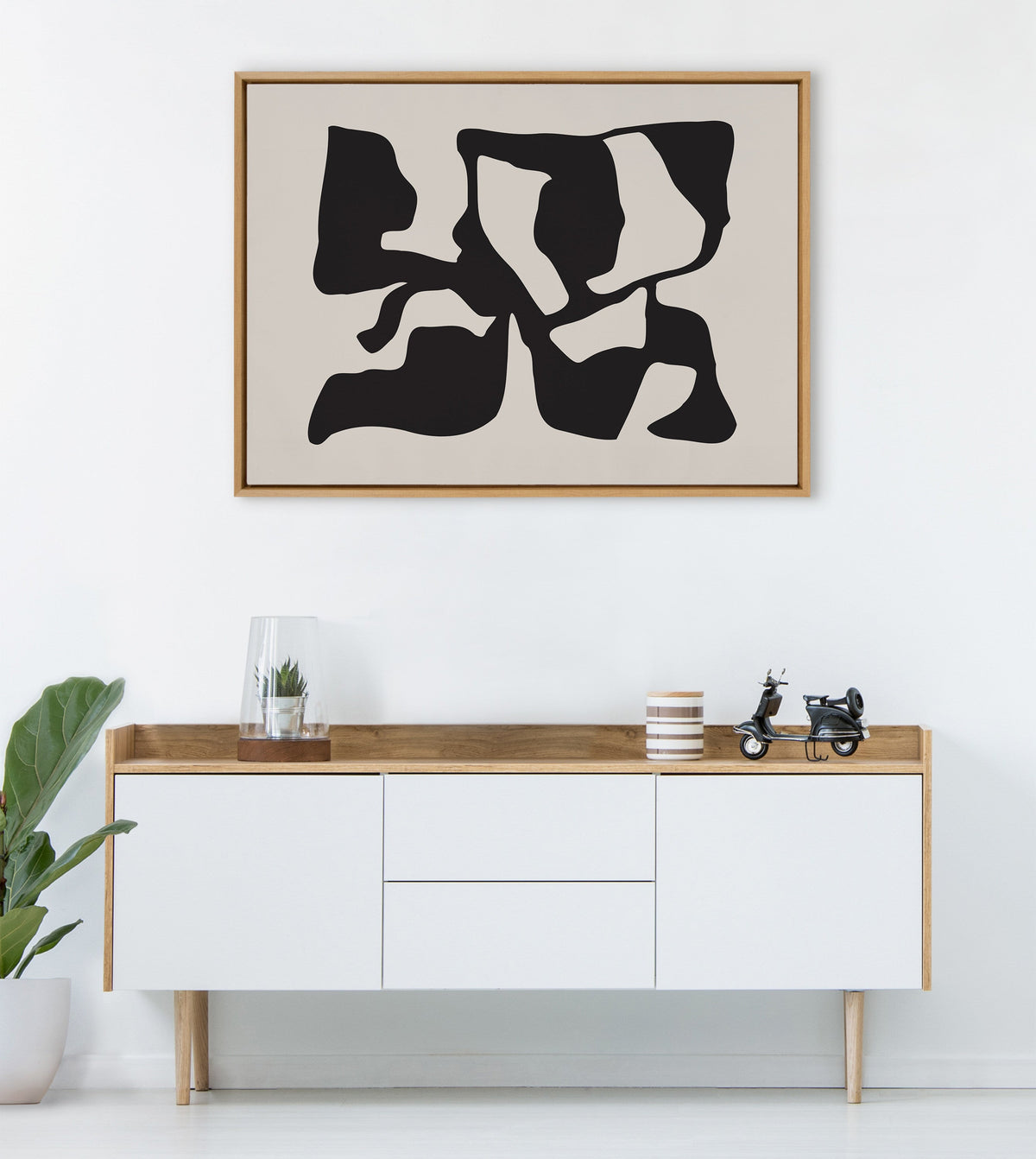 Abstract black distorted shapes on tan background framed in simple wood, hanging above a minimalist white and wood sideboard with a plant, glass terrarium, striped cup, and toy scooter.